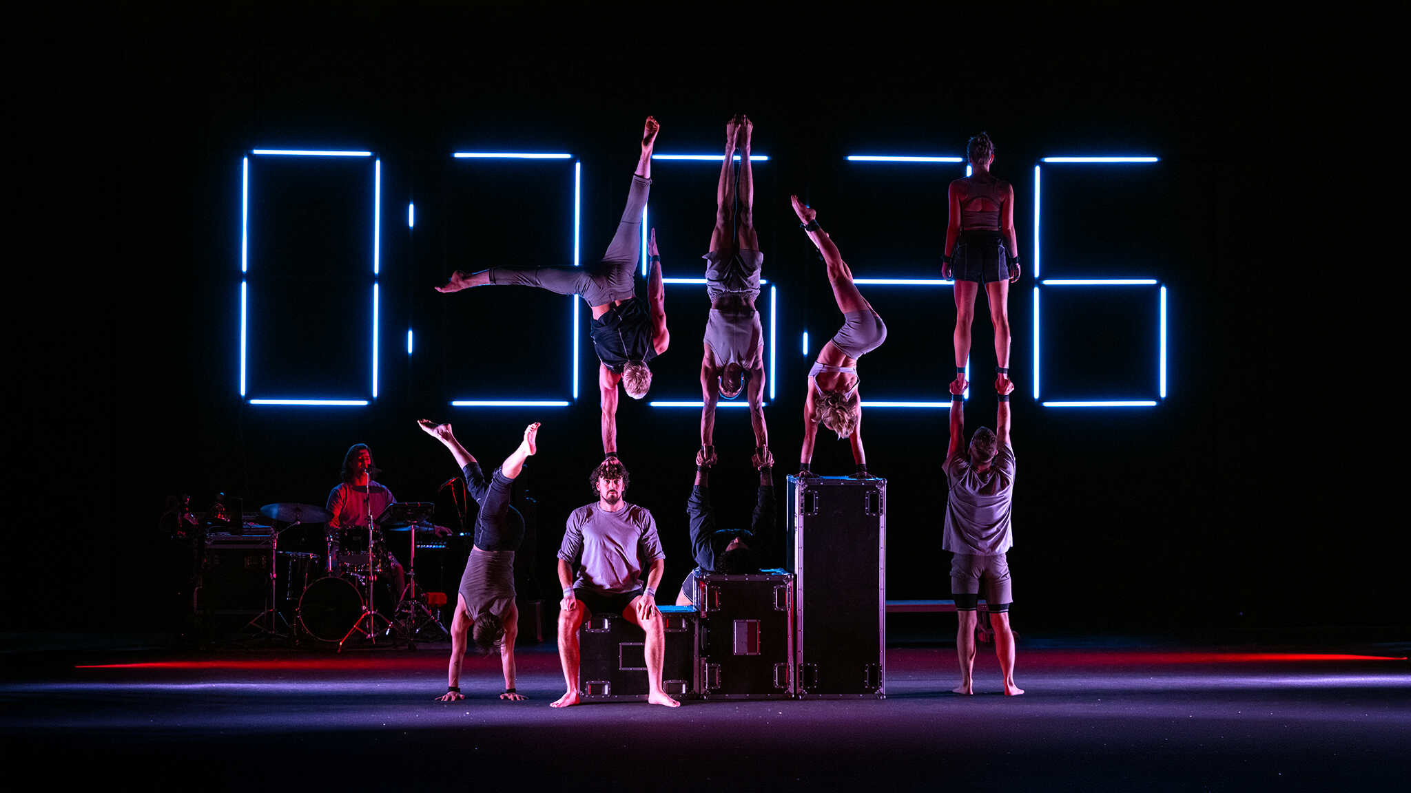 8 acrobats are seen on stage, half are balancing on other's heads while one is seen playing the drums, a large timer is seen behind them all